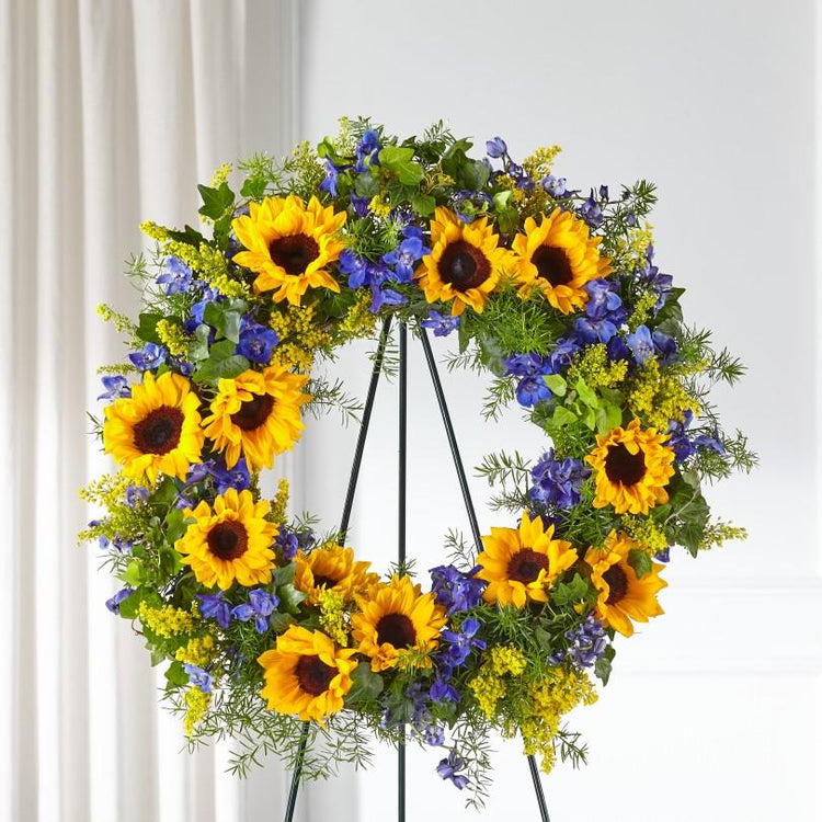 blue delpihinium, sunflowers, solidago, and lush greens arranged in a funeral wreath. Wreath is approximately 26" in diameter